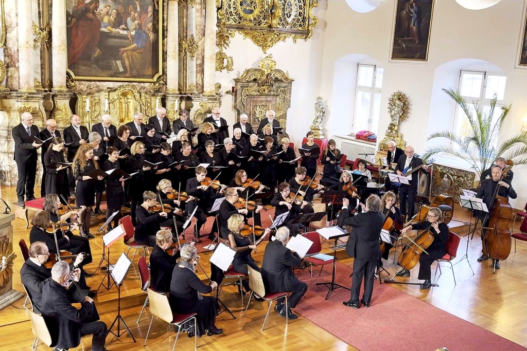 Amberger Chorgemeinschaft und Orchester der OTH Amberg-Weiden in Konzertkleidung während des Konzerts im Kongregationssaal St. Georg, aufgestellt bzw. sitzend vor dem barocken Hochaltar.